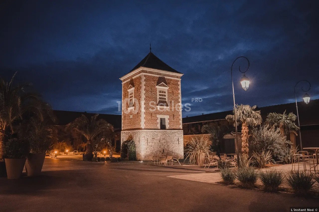 Tour en briques éclairée la nuit, entourée de palmiers et de lampadaires dans une cour pavée.