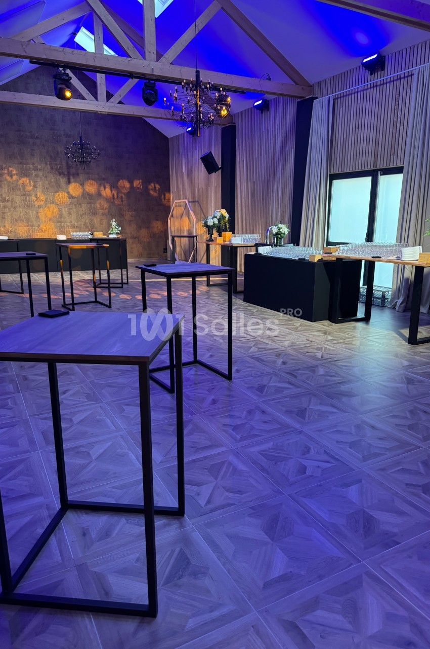 Salle de réception moderne avec tables hautes, décoration élégante, éclairage bleu et grandes fenêtres.