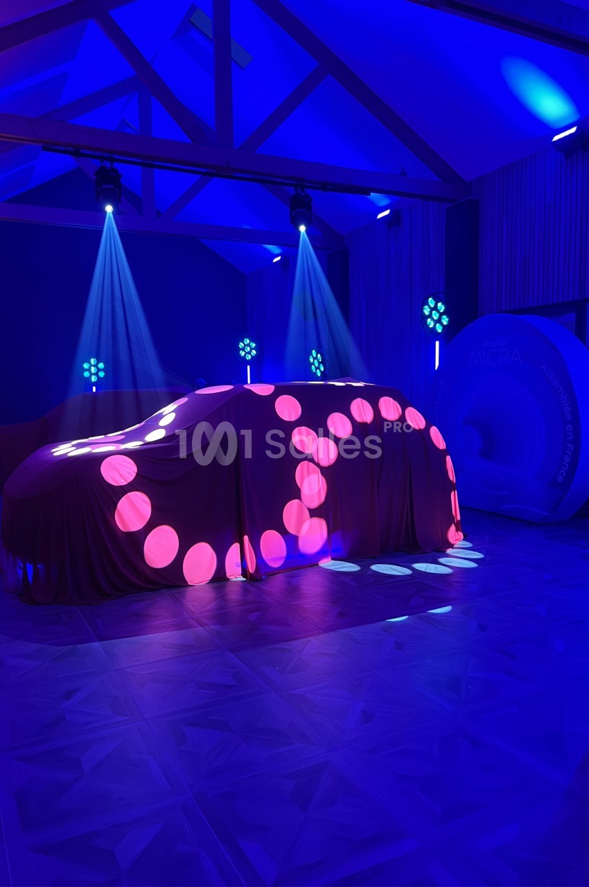 Voiture recouverte d'un drap illuminé par des motifs lumineux roses et bleus dans une salle éclairée en bleu.