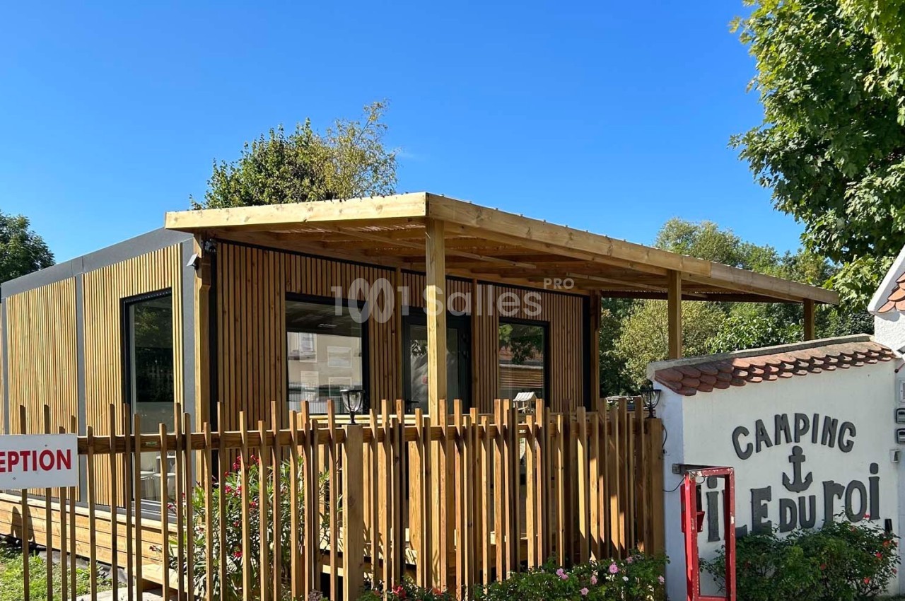 Bâtiment en bois moderne avec terrasse couverte, situé à l'entrée d'un camping, entouré de verdure.