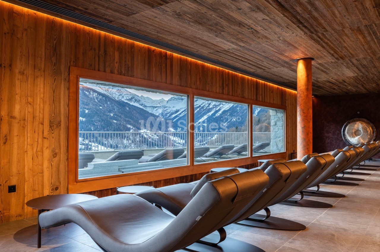 Salle de repos avec chaises longues orientées vers une grande baie vitrée offrant une vue sur des montagnes enneigées.