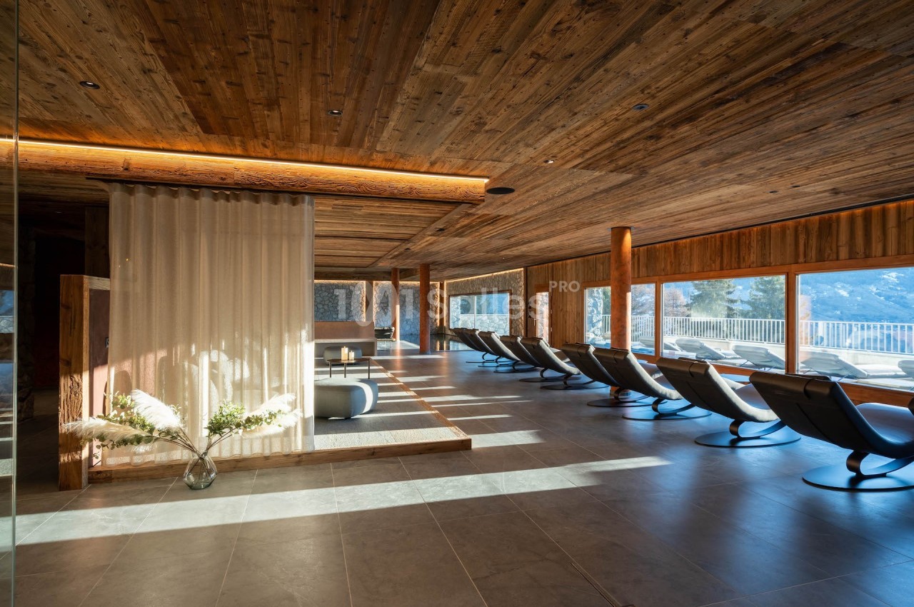 Salle de relaxation lumineuse avec chaises longues, plafond en bois et grandes baies vitrées offrant une vue sur l'extérieur.