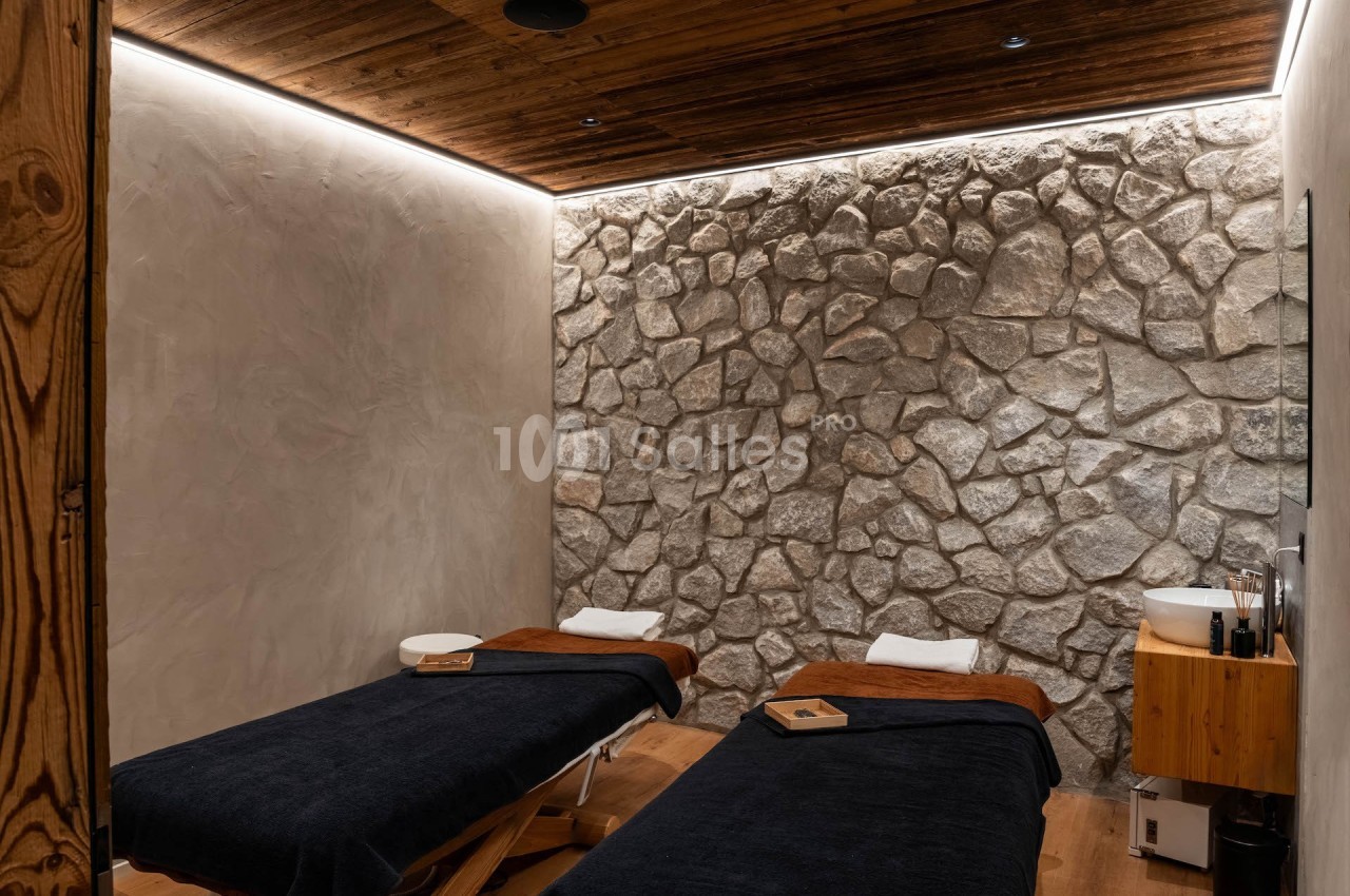 Salle de soins avec deux tables de massage, murs en pierre, éclairage tamisé et ambiance chaleureuse.