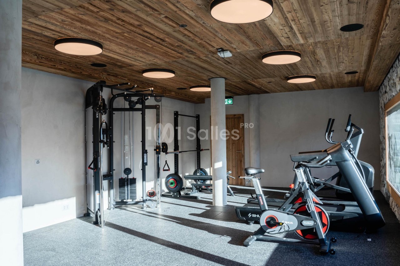 Salle de sport lumineuse avec équipements de musculation, tapis de course et vélo d'entraînement, plafond en bois.