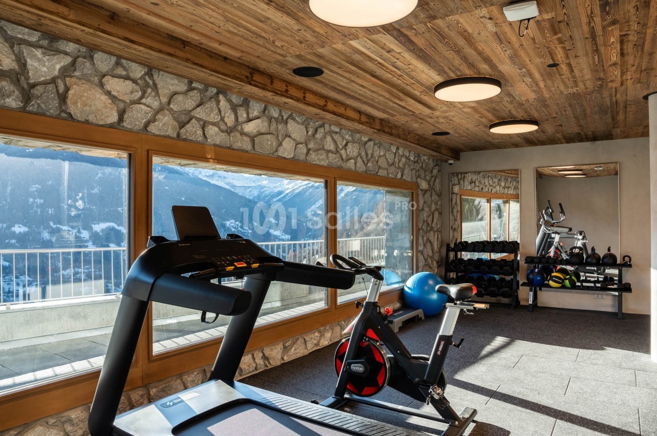 Salle de sport lumineuse avec tapis de course, vélo d'appartement, haltères et vue sur les montagnes à travers de grandes…