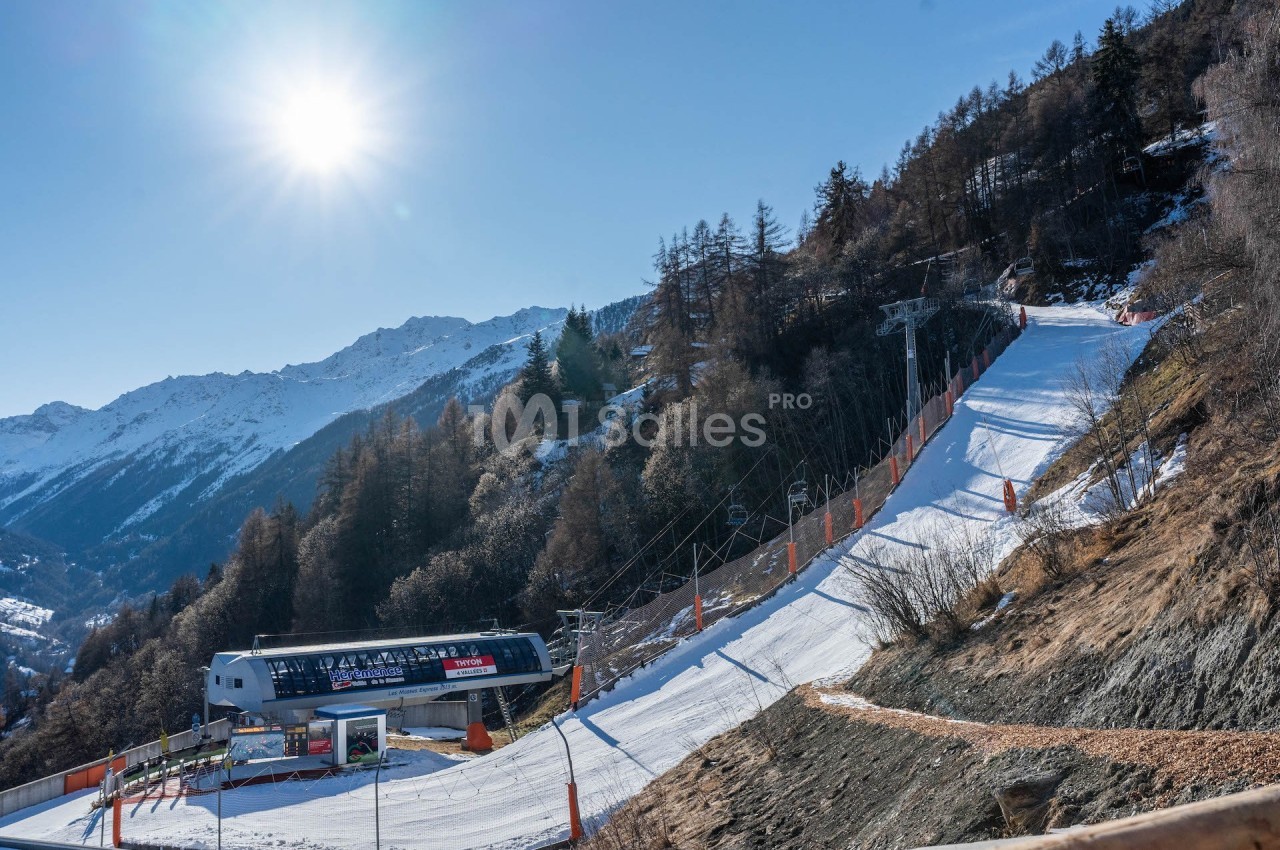 Piste de ski ensoleillée bordée de sapins avec un télésiège et des montagnes en arrière-plan.