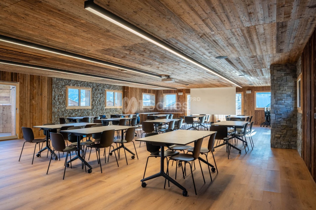 Salle de réunion lumineuse avec tables et chaises modernes, murs en bois et pierre, et écran de projection.
