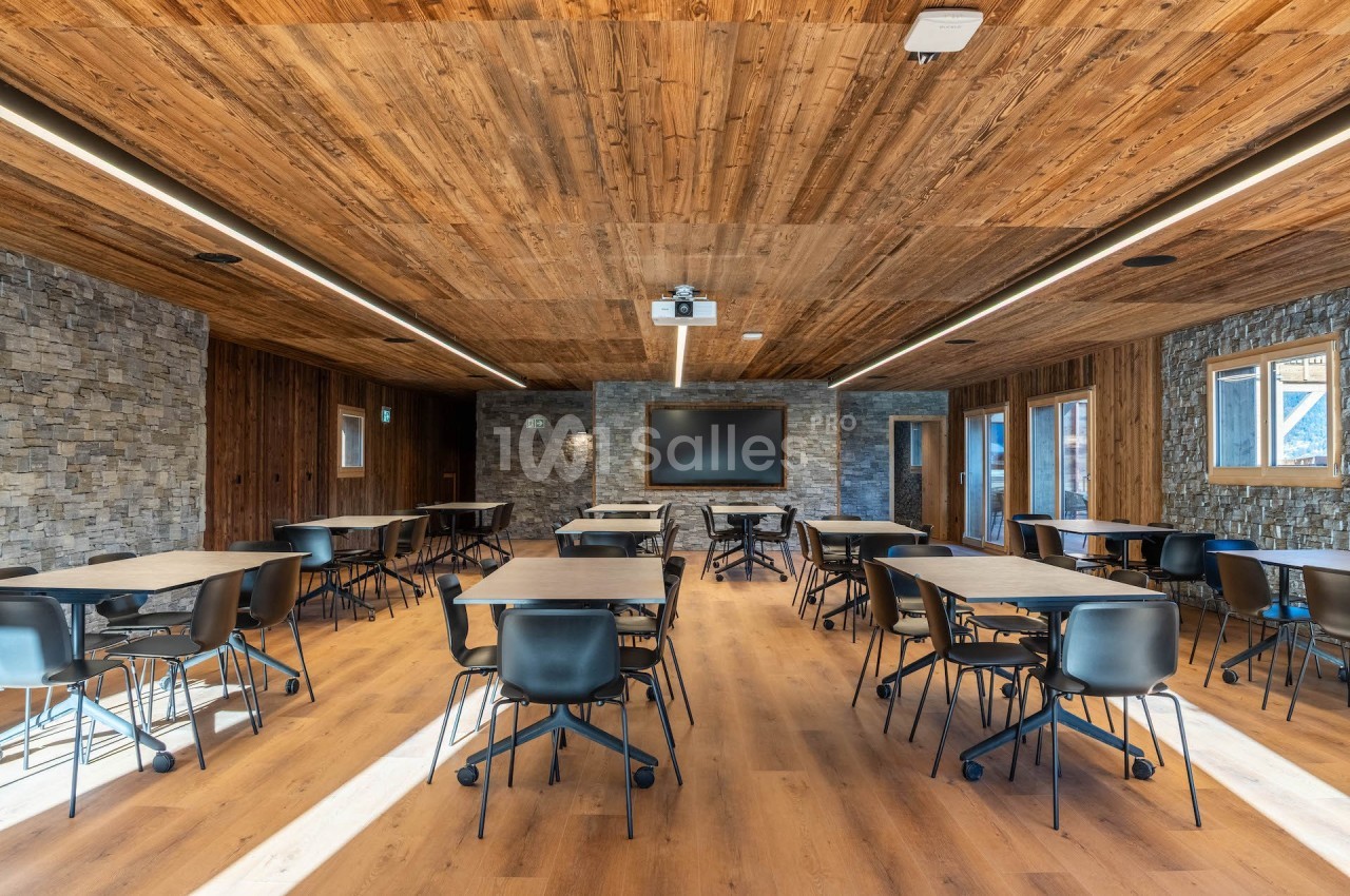 Salle de réunion moderne avec murs en pierre, plafond en bois, tables et chaises noires disposées en rangées.