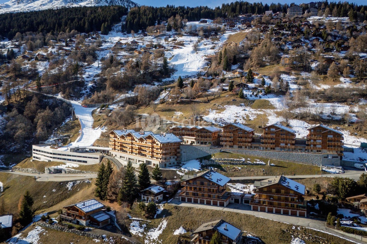Vue aérienne d'une station de montagne avec chalets en bois, pistes de ski et forêt en arrière-plan.