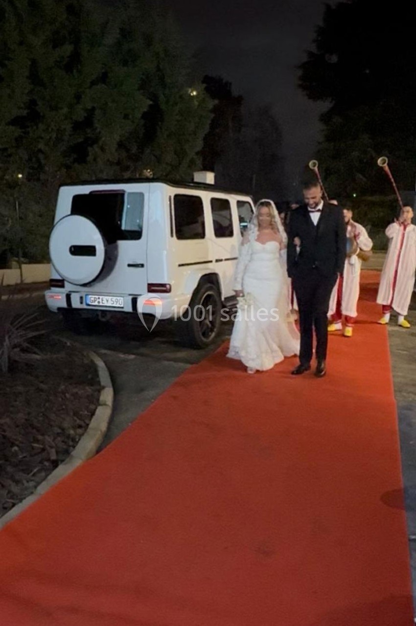 Un couple en tenue de mariage marche sur un tapis rouge, suivi par des personnes en habits religieux, près d'un 4x4 blanc.
