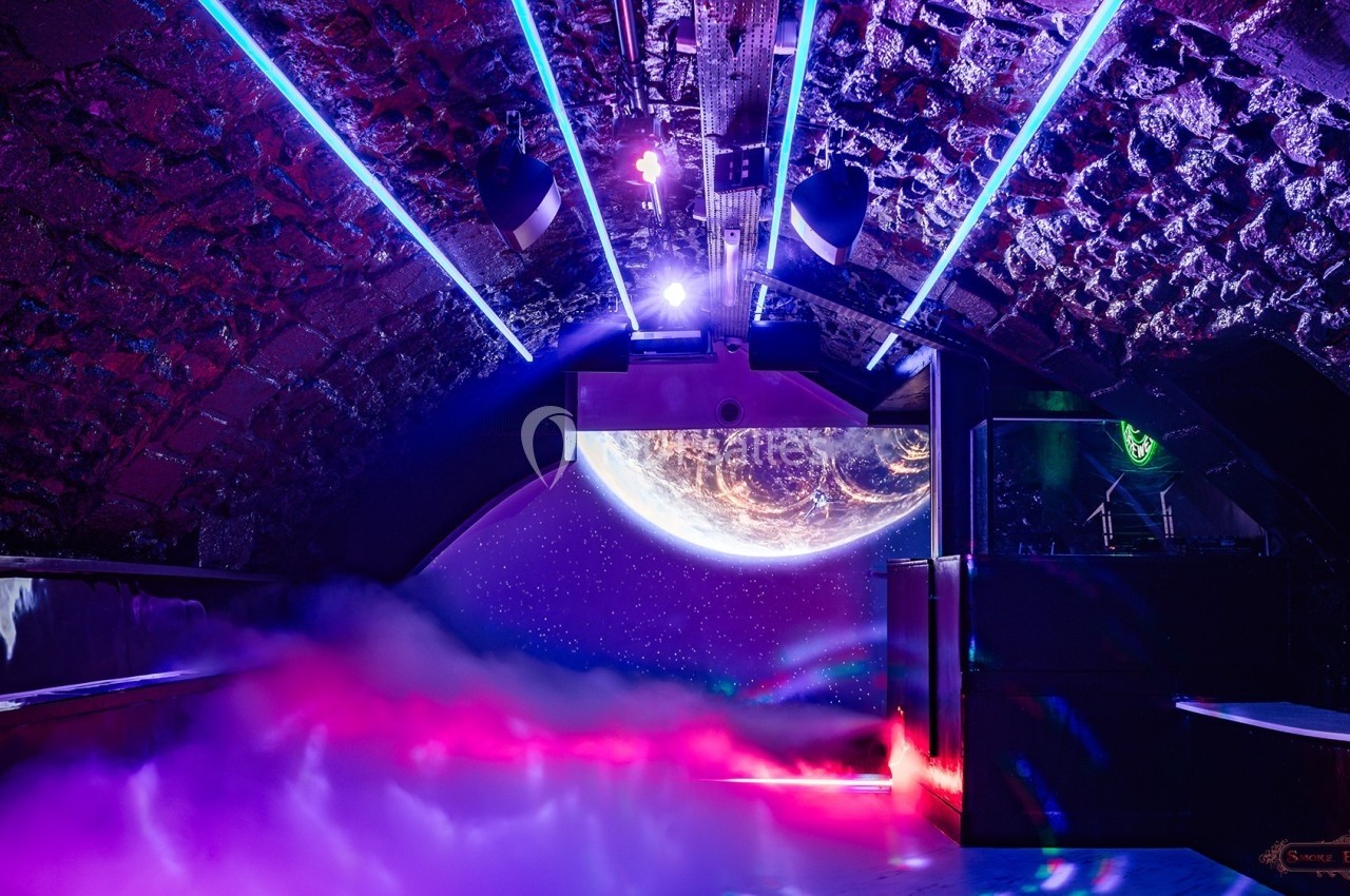 Salle voûtée en pierre avec éclairage néon coloré, fumée au sol et projection d'une planète en arrière-plan.