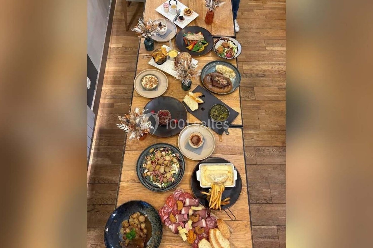 Table en bois dressée avec divers plats, incluant charcuterie, fromage fondu, salades et desserts, dans un cadre chaleureux.