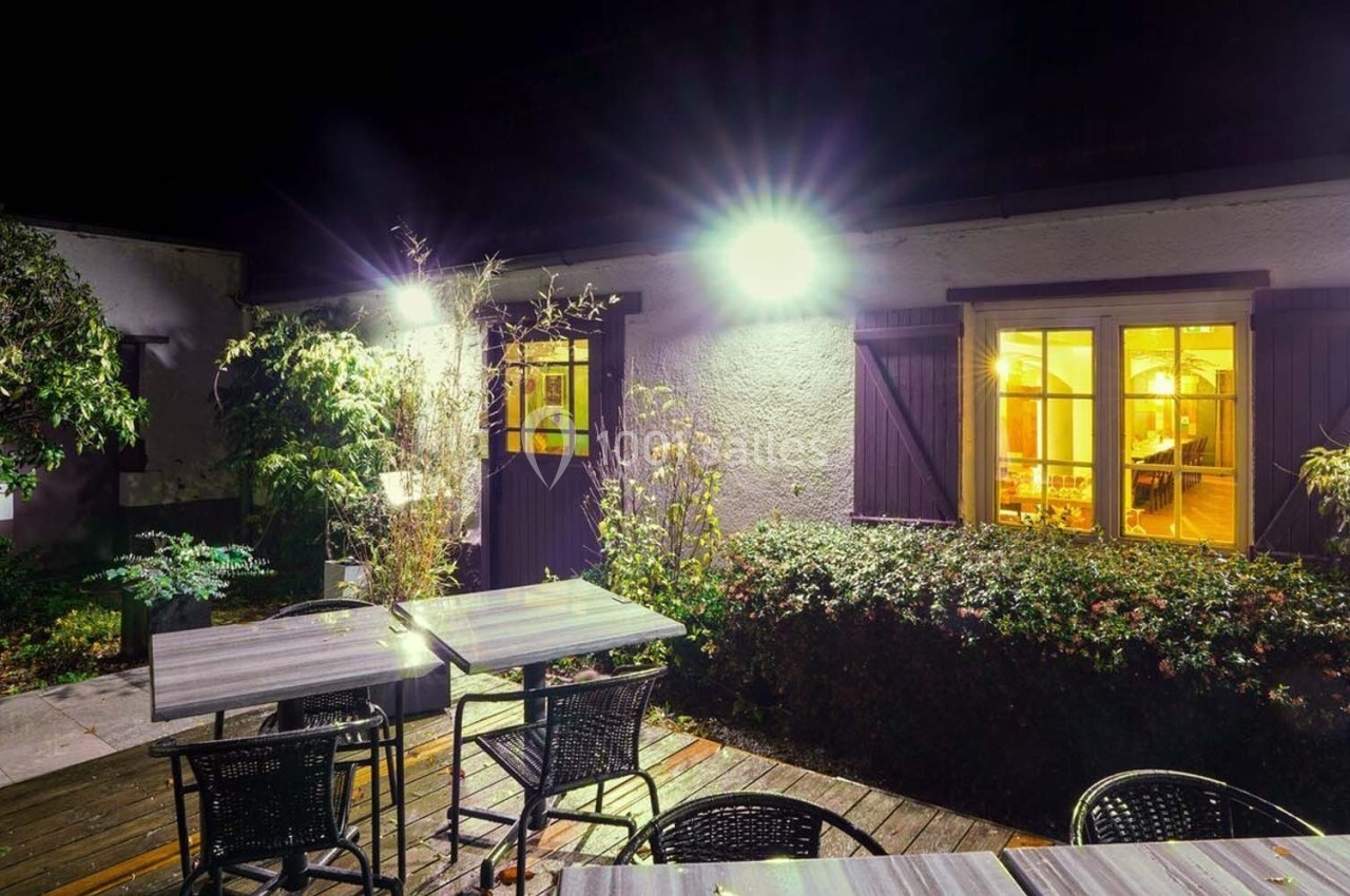 Terrasse extérieure éclairée la nuit avec tables, chaises et vue sur une maison aux fenêtres illuminées.