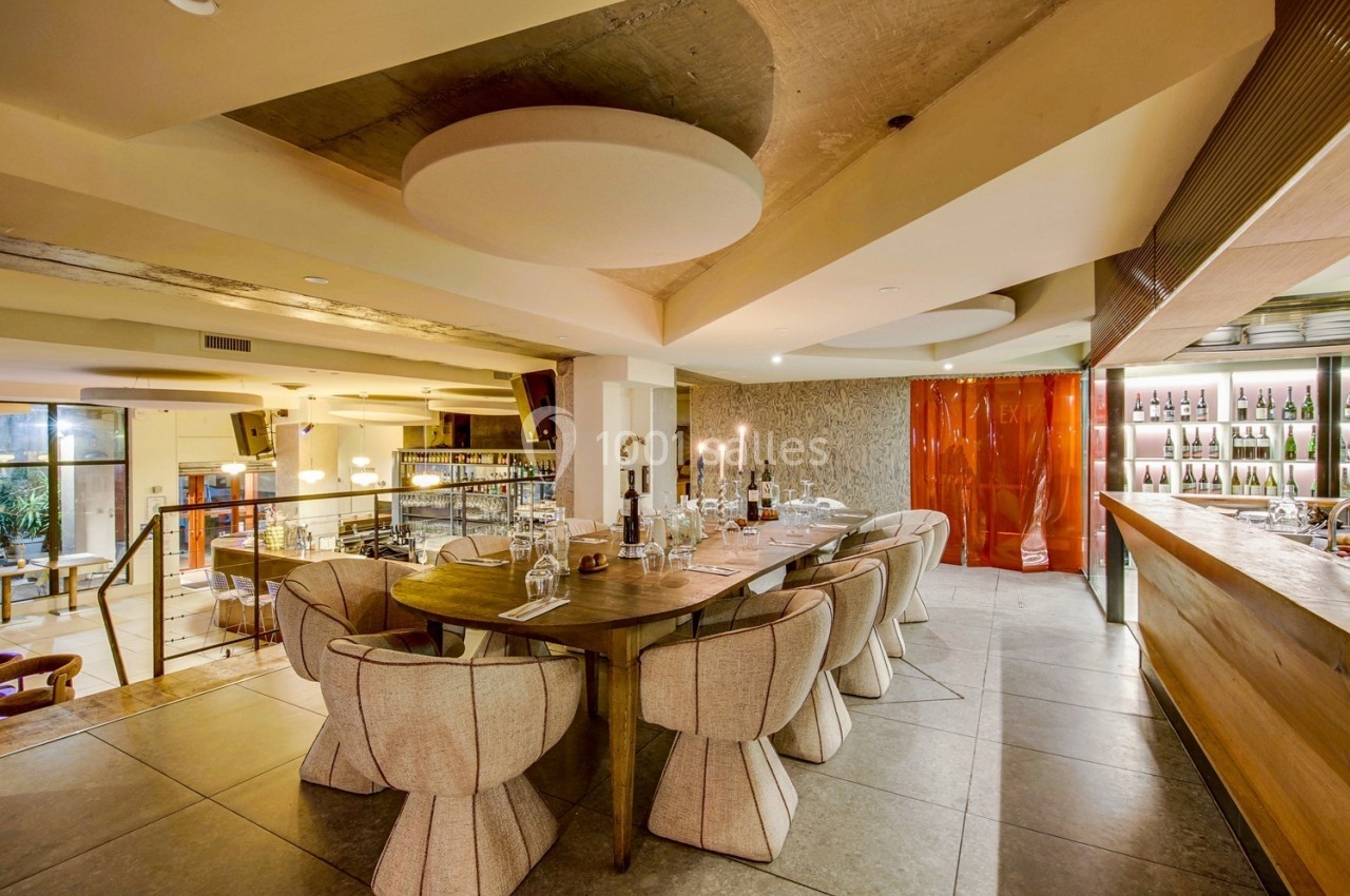 Salle de restaurant moderne avec grande table en bois, chaises design, éclairage chaleureux et bar en arrière-plan.