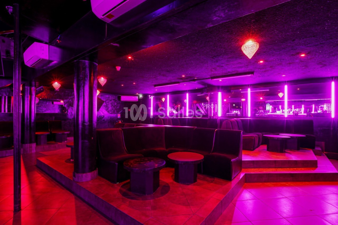 Salle de bar ou club avec éclairage néon rose, banquettes noires, tables basses et comptoir en arrière-plan.