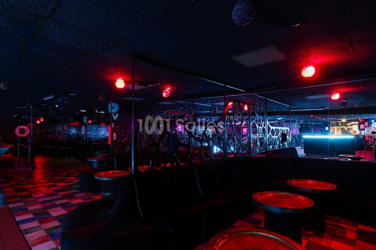 Salle de club avec éclairage rouge tamisé, banquettes noires, tables rondes et ambiance nocturne.