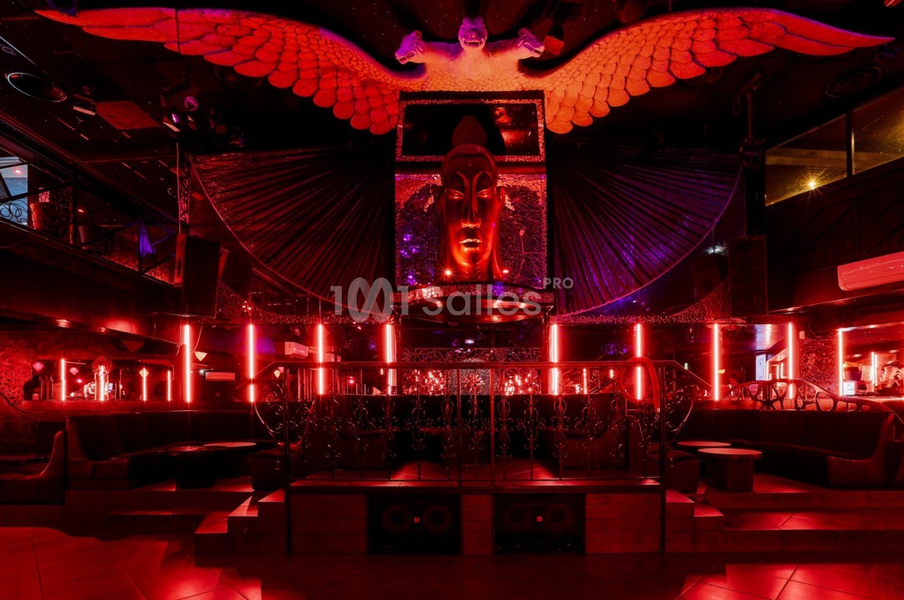 Salle de club avec éclairage rouge, décorations sombres, statue centrale et ailes illuminées en arrière-plan.