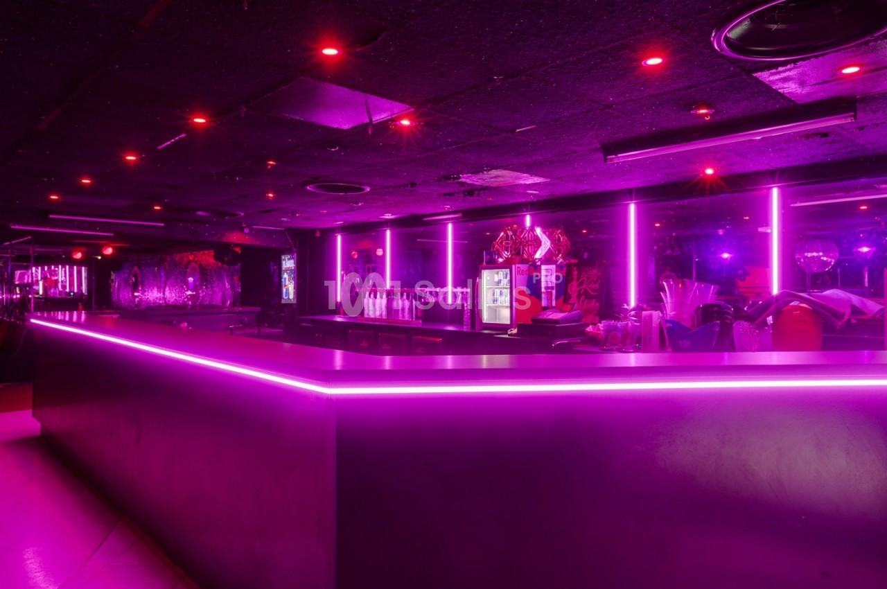 Bar avec éclairage néon rose, comptoir lumineux et rangées de bouteilles en arrière-plan dans une ambiance sombre.