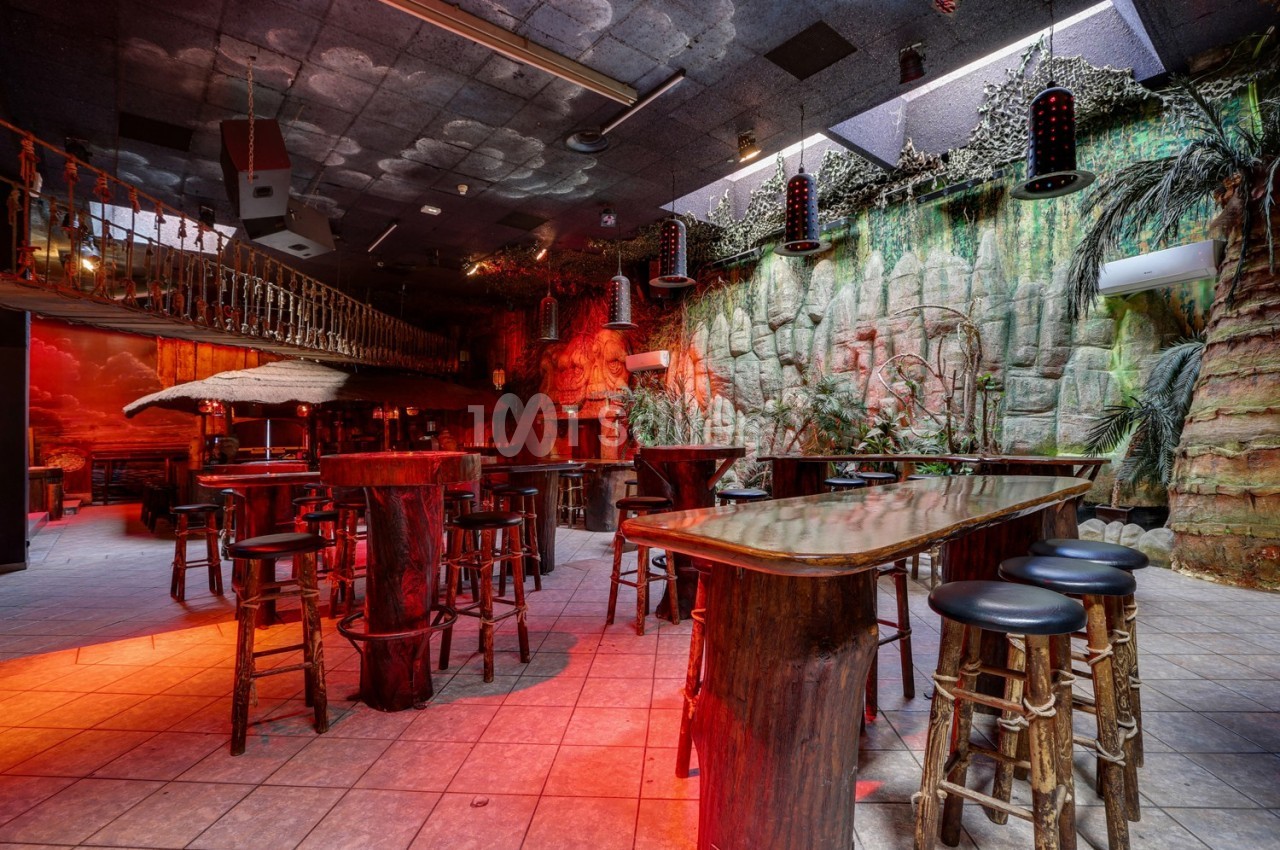 Salle de bar à thème tropical avec tables et tabourets en bois, éclairage rouge et décor végétal exotique.
