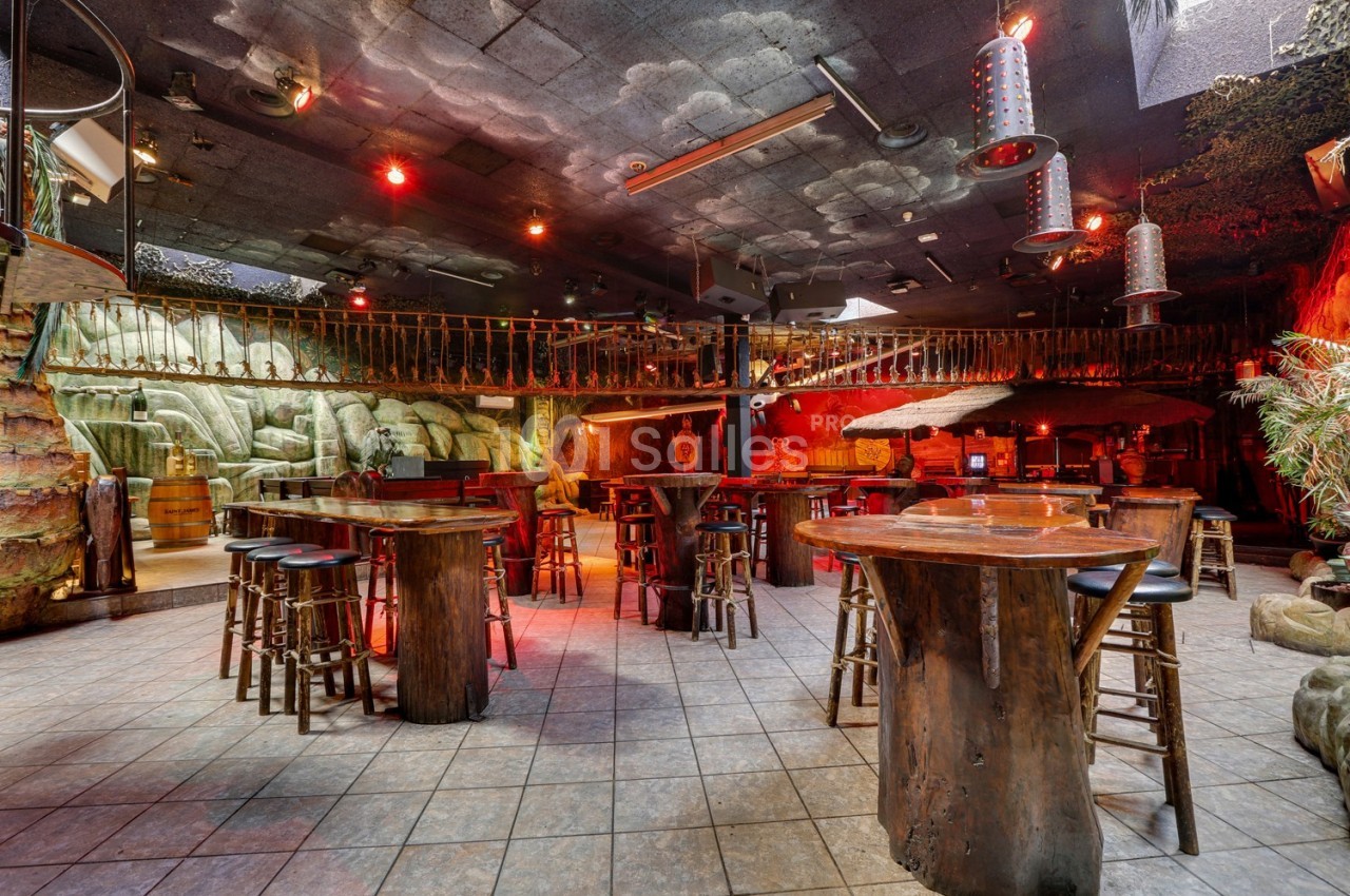 Salle de bar à thème avec tables en bois, décor tropical, éclairage tamisé et mezzanine en arrière-plan.