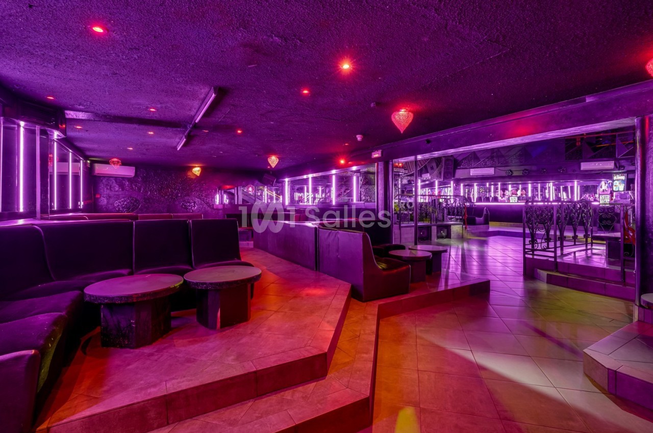 Intérieur d'un club avec éclairage violet, banquettes, tables basses et sol carrelé.