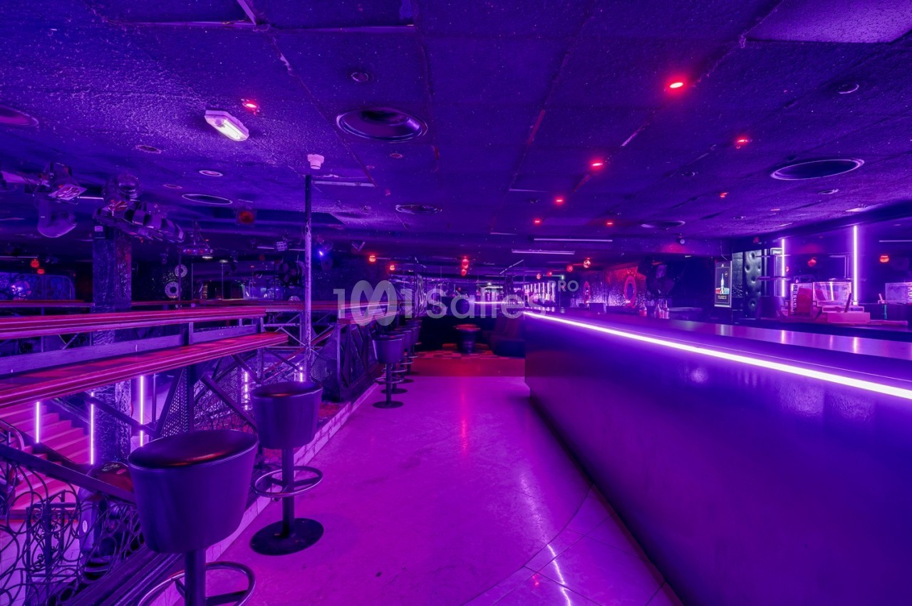 Salle de discothèque vide avec éclairage violet, comptoir lumineux et tabourets alignés le long d'une rambarde.