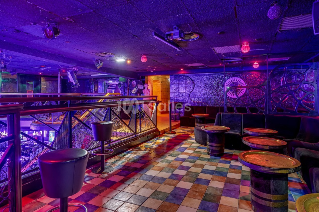 Salle de bar colorée avec tables rondes, sièges en cuir noir et éclairage violet dans une ambiance nocturne.