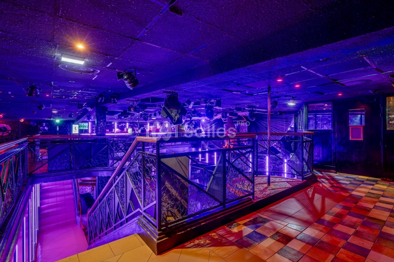 Intérieur d'une discothèque avec éclairage violet, escalier central, balustrades métalliques et sol carrelé coloré.