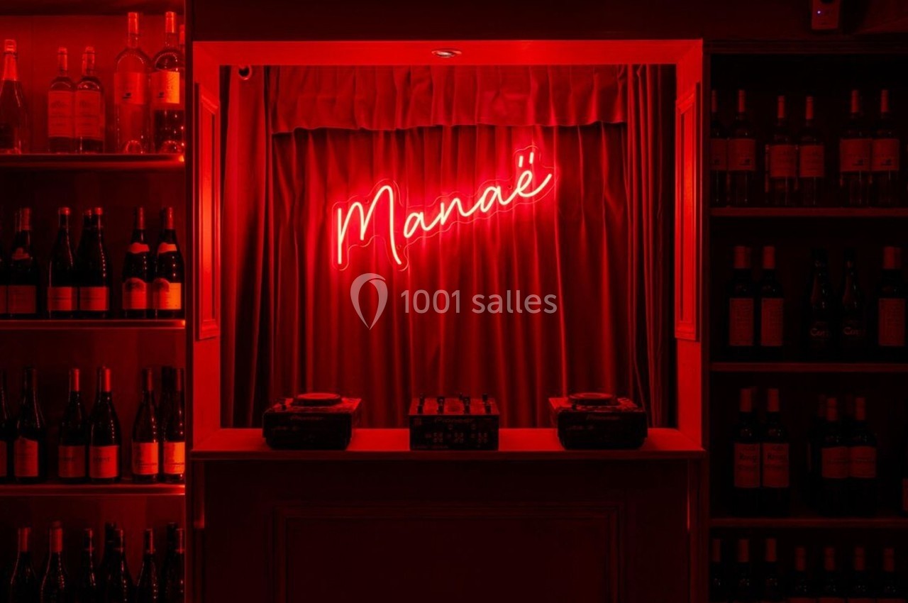 Enseigne lumineuse rouge ’Manaé’ au-dessus d'un comptoir, entourée d'étagères remplies de bouteilles de vin.