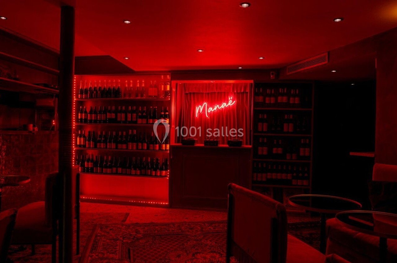 Bar à l'éclairage rouge tamisé avec étagères de bouteilles, enseigne néon ’Monaie’ et mobilier sombre.