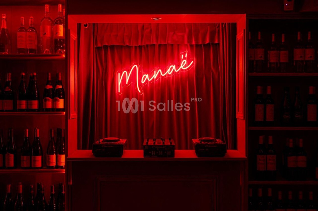 Étalage de bouteilles de vin entourant un comptoir éclairé par une enseigne néon rouge indiquant ’Manaé’.