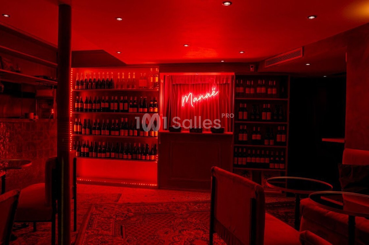 Bar à l'éclairage rouge tamisé avec étagères de bouteilles, tables basses et fauteuils dans une ambiance chaleureuse.