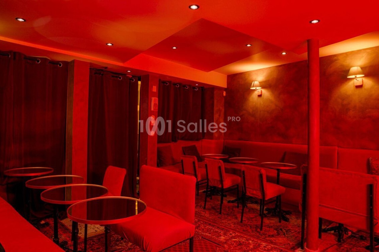 Salle intime éclairée de lumière rouge, avec des tables rondes, des chaises rembourrées et des rideaux sombres.