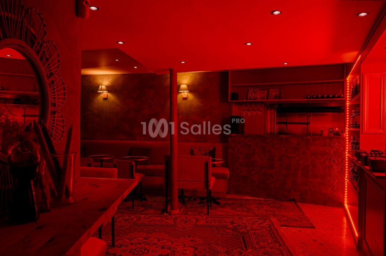 Salle intimiste éclairée par une lumière rouge, avec des fauteuils, un bar et des étagères de bouteilles.