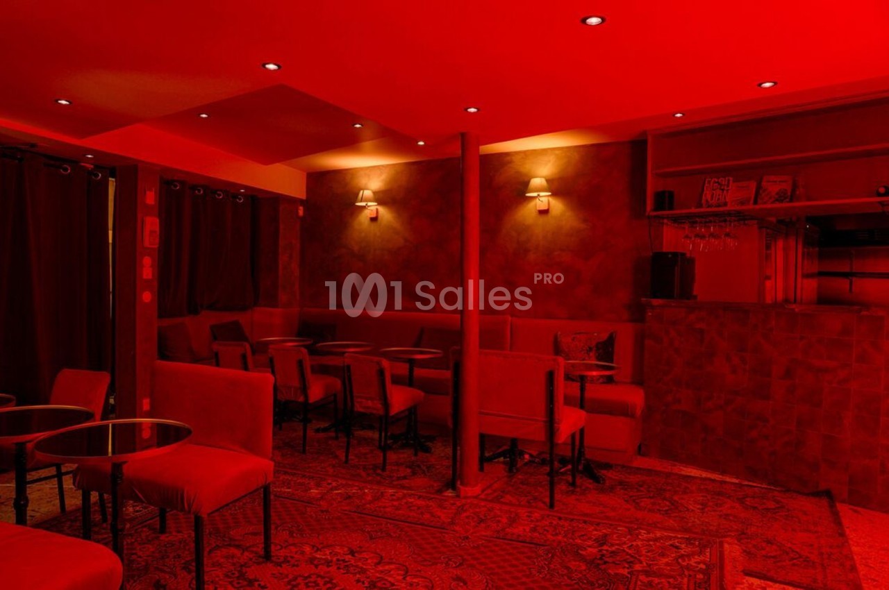 Salle de bar intimiste éclairée de lumières rouges, avec tables, chaises et comptoir en arrière-plan.
