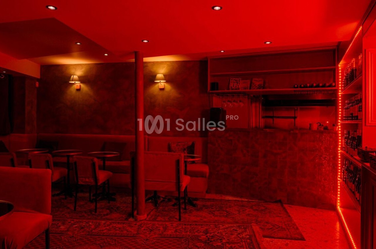 Salle de bar à l'éclairage rouge, avec tables, chaises, comptoir et étagères garnies de bouteilles.