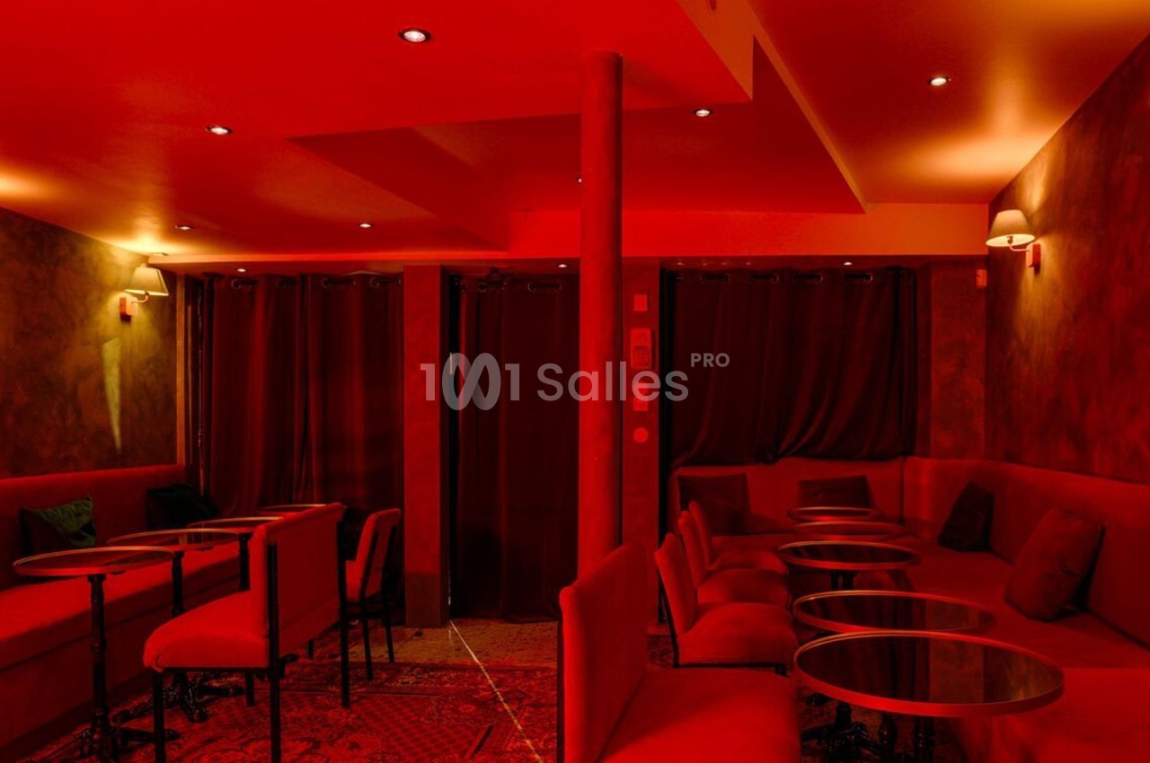 Salle intimiste éclairée de lumières rouges, avec canapés, petites tables rondes et rideaux sombres en arrière-plan.