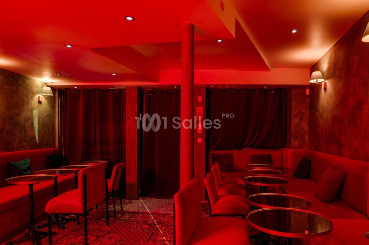 Salle intimiste éclairée par une lumière rouge, avec banquettes, petites tables rondes et rideaux sombres en arrière-plan.