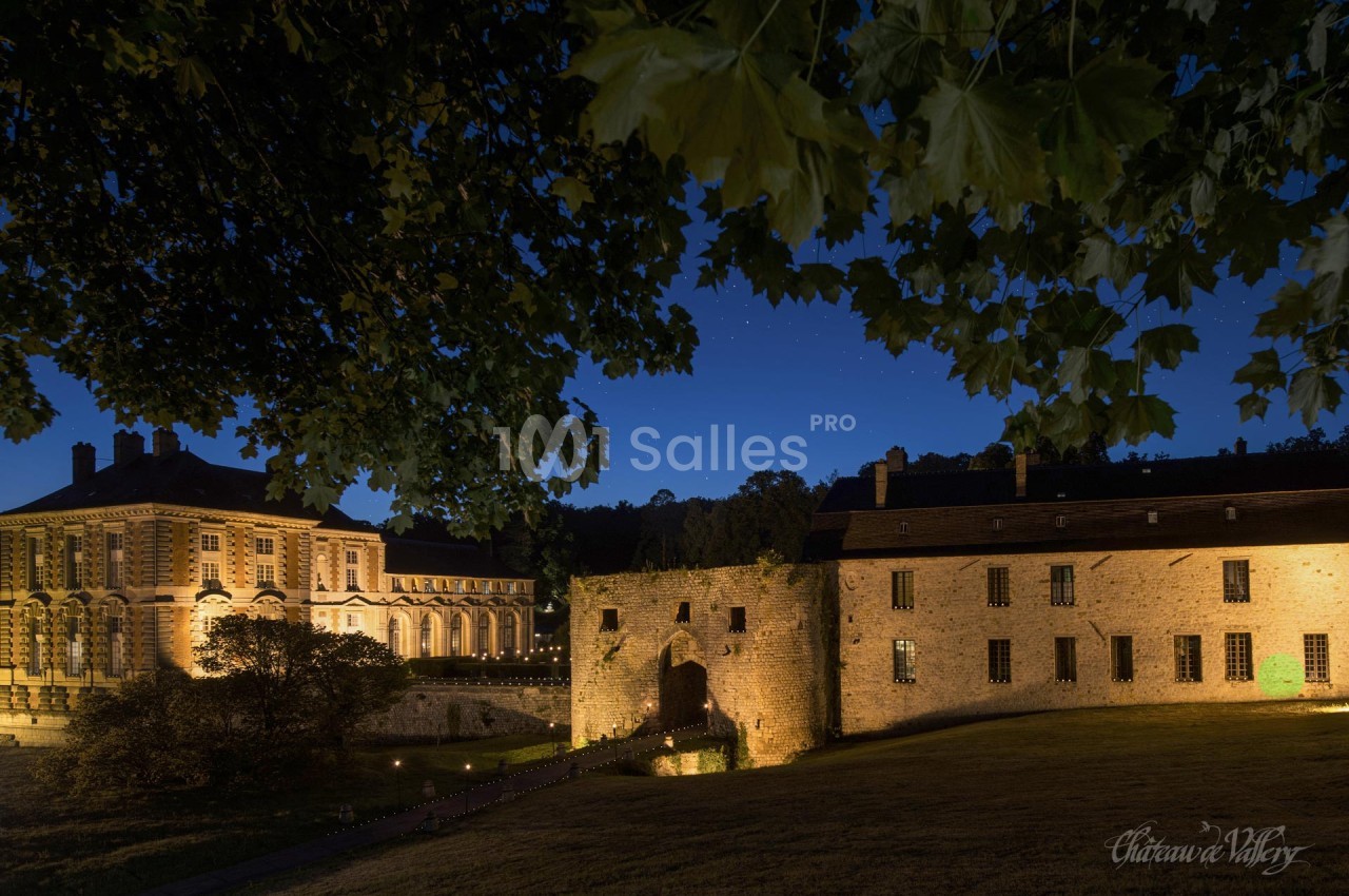 Vue nocturne d'un château éclairé avec des bâtiments en pierre et un portail entouré de verdure.