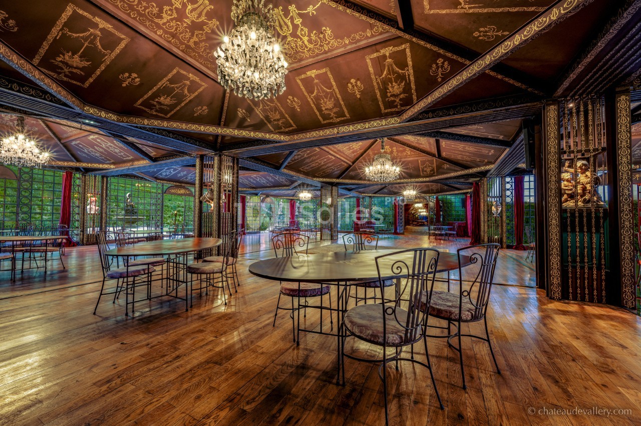 Salle spacieuse avec parquet en bois, tables et chaises en fer forgé, lustres suspendus et décorations murales élégantes.