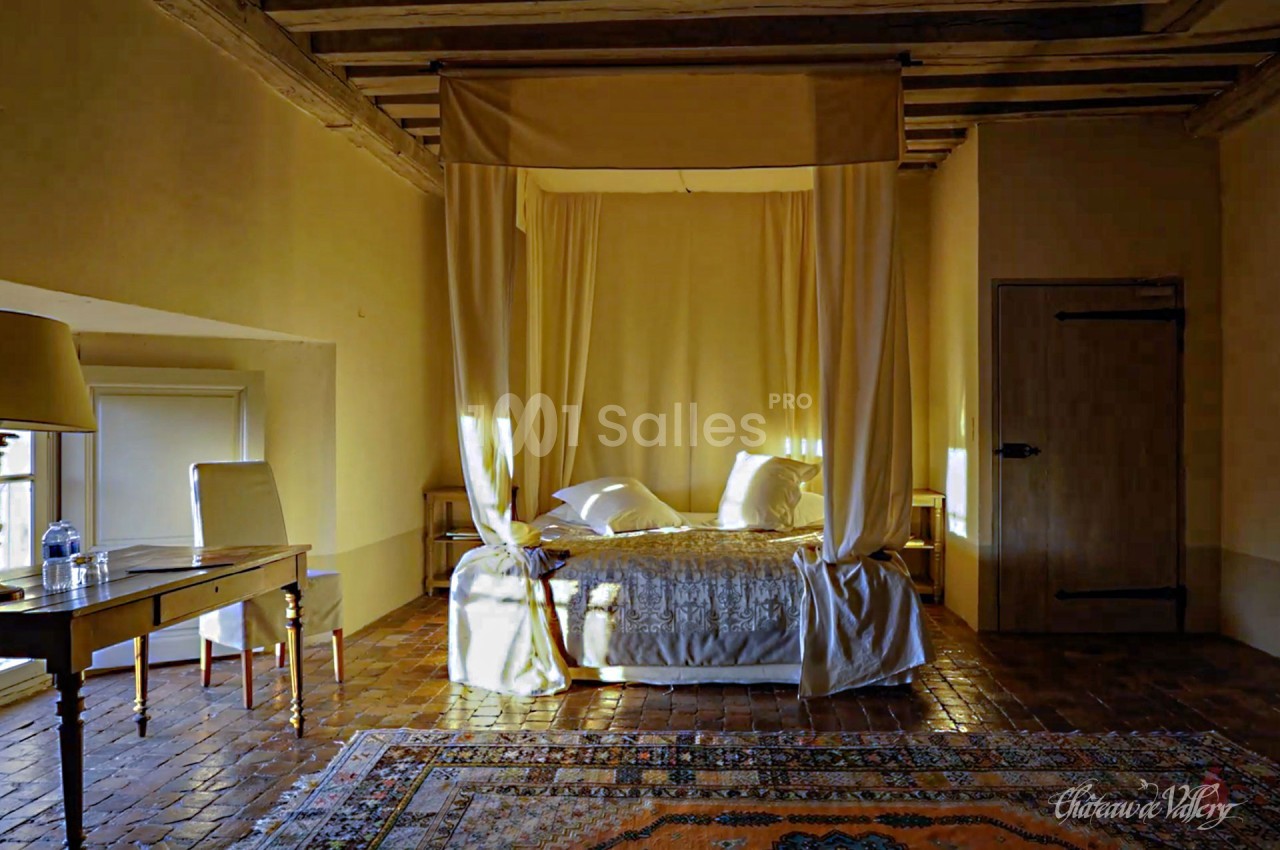 Lit à baldaquin dans une chambre rustique avec murs jaunes, sol en terre cuite et tapis coloré.