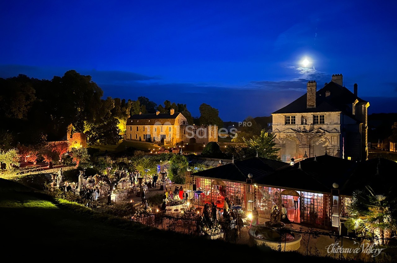 Vue nocturne d'un château éclairé avec des invités rassemblés dans un jardin illuminé sous une pleine lune.