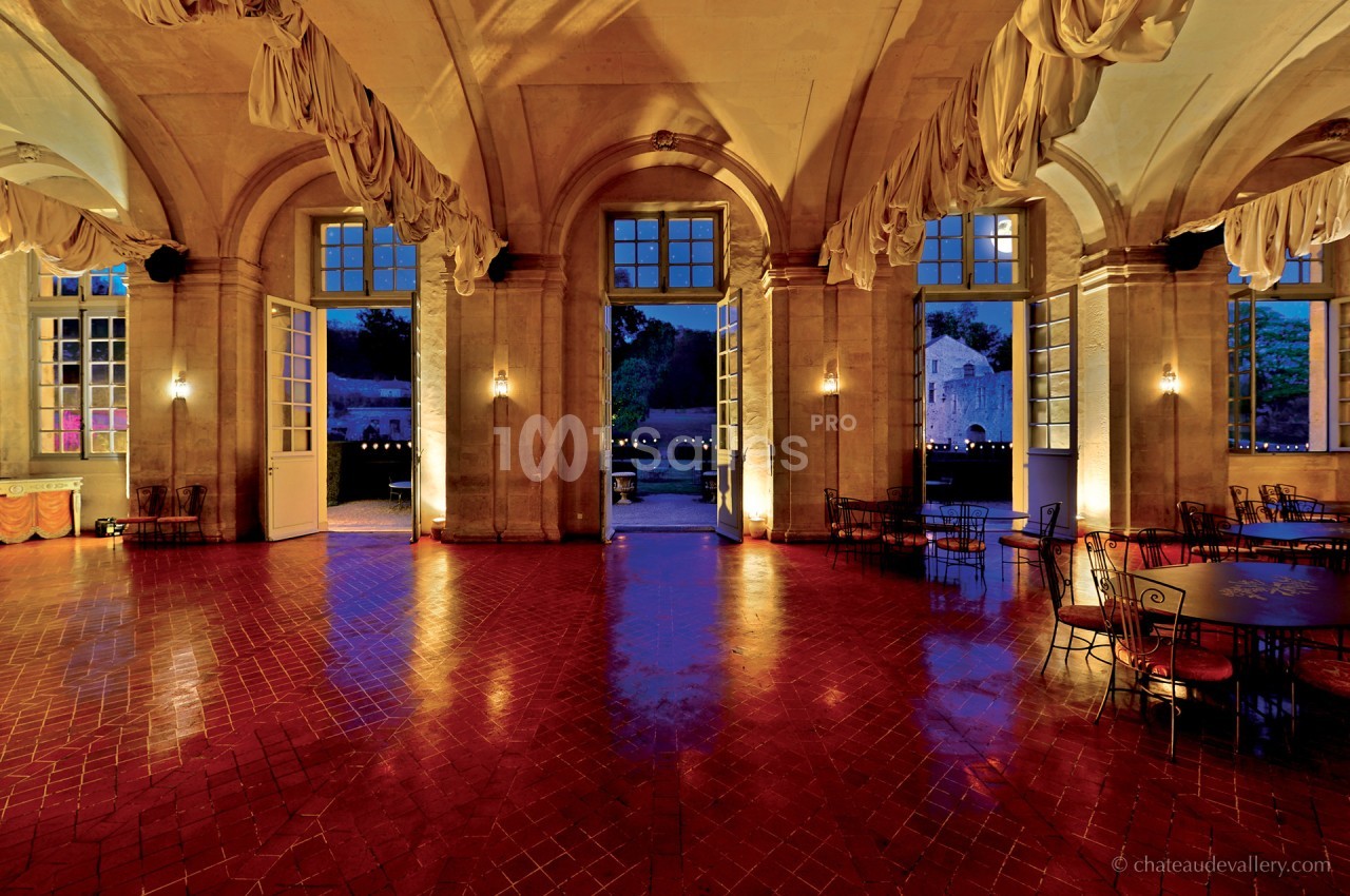 Salle spacieuse avec sol en carreaux rouges, grandes arches vitrées ouvertes sur un jardin éclairé au crépuscule.