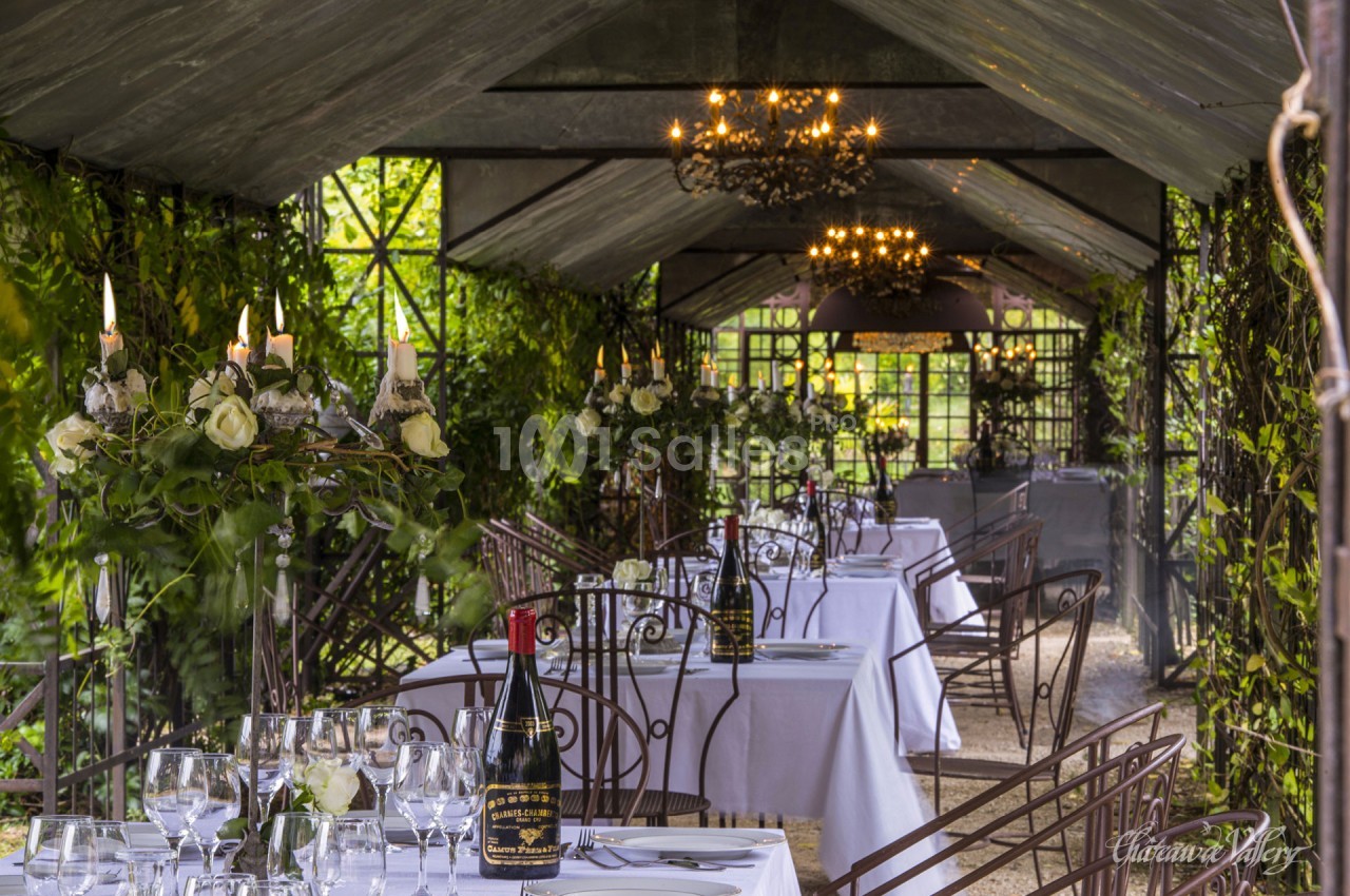 Salle à manger extérieure couverte, décorée de verdure, avec tables dressées et chandeliers suspendus.
