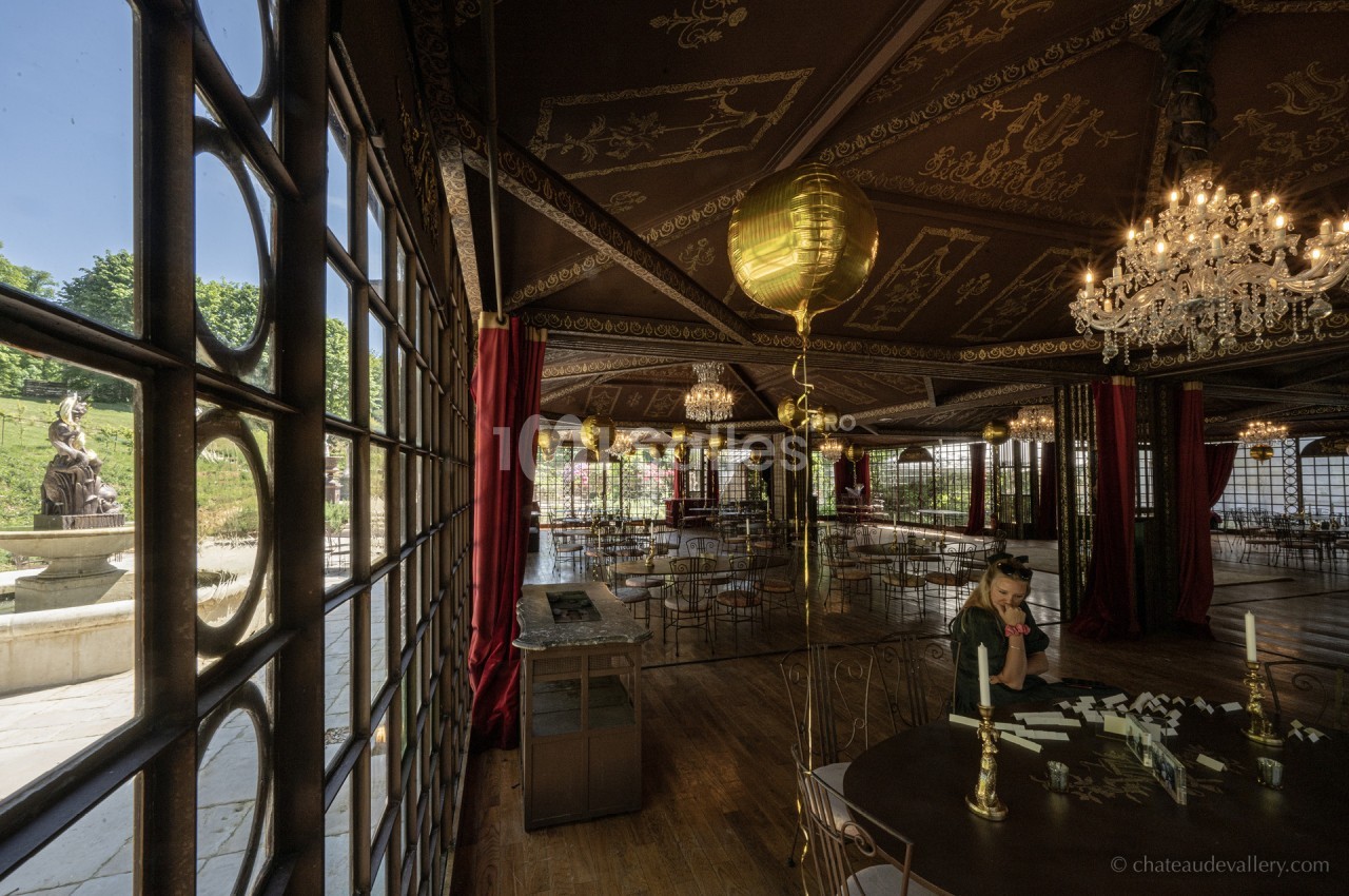 Salle élégante avec grandes baies vitrées, lustres en cristal, décor doré et vue sur une statue dans un jardin.