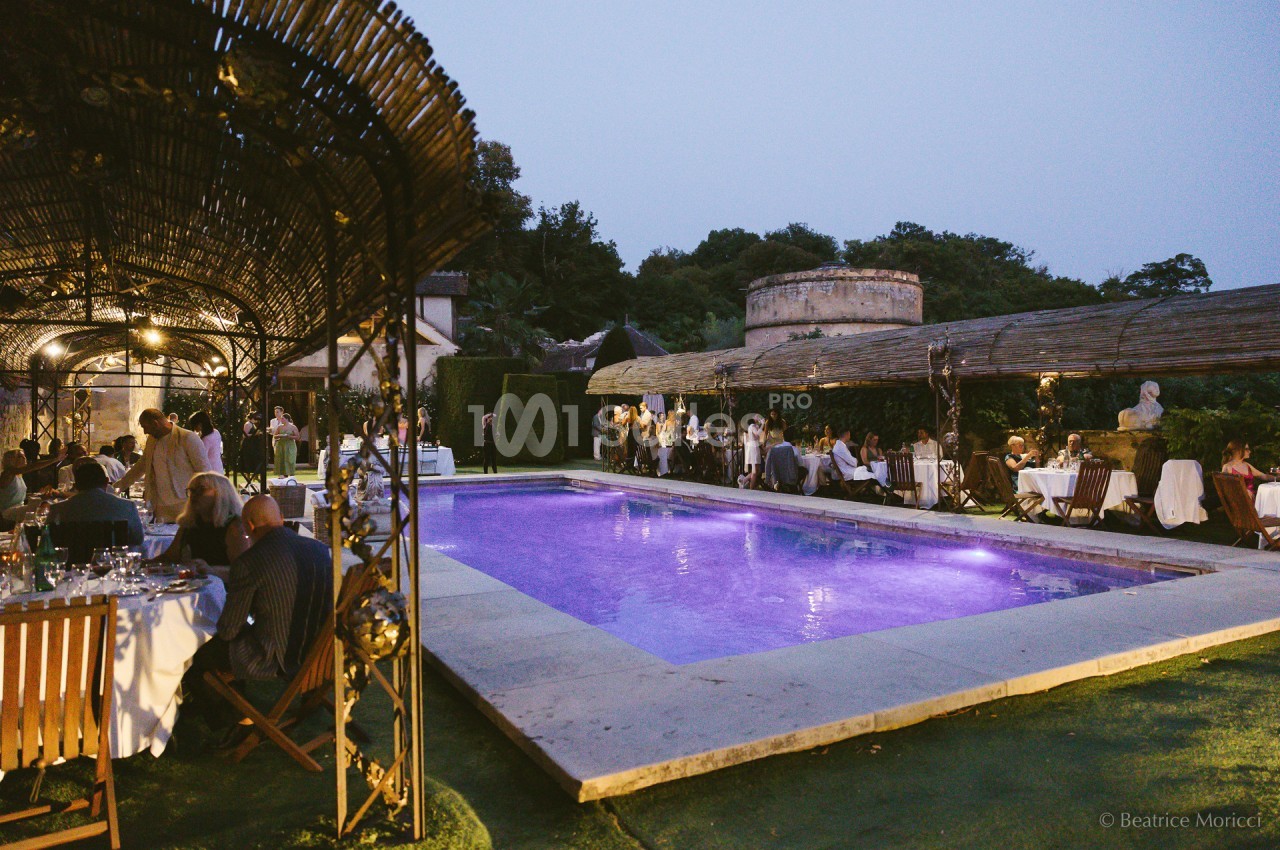 Dîner en plein air autour d'une piscine éclairée, avec des invités assis sous des pergolas au crépuscule.