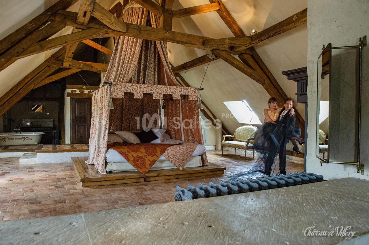 Chambre rustique avec lit à baldaquin, poutres apparentes et couple debout près d'une fenêtre.