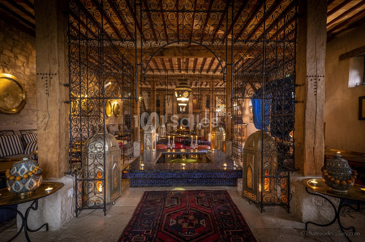 Intérieur d'une salle voûtée avec décorations orientales, lanternes, tapis et bassin central entouré de grilles en fer forgé.