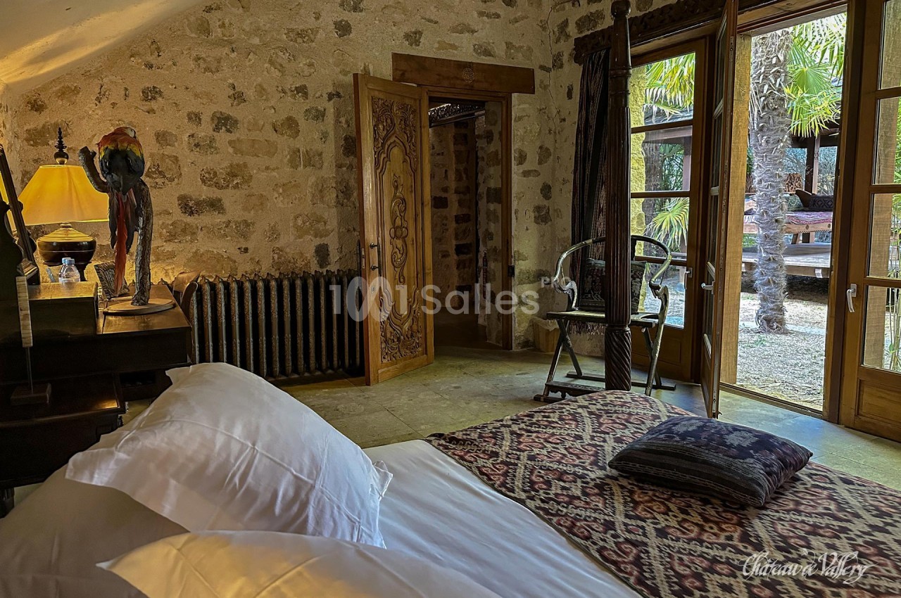 Chambre avec lit, tapis décoratif, mobilier en bois, et vue sur un jardin avec palmiers à travers des portes vitrées.