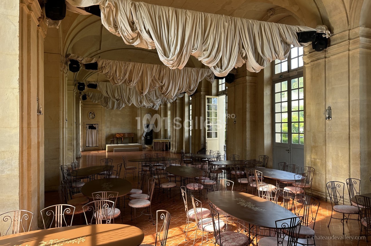Salle lumineuse avec tables et chaises en fer forgé, décorée de drapés suspendus au plafond et grandes fenêtres vitrées.